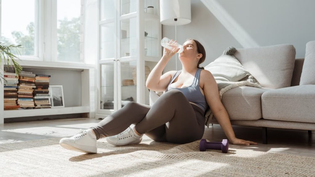 Woman sitting on living room floor, drinking water after workout, enjoying relaxation.