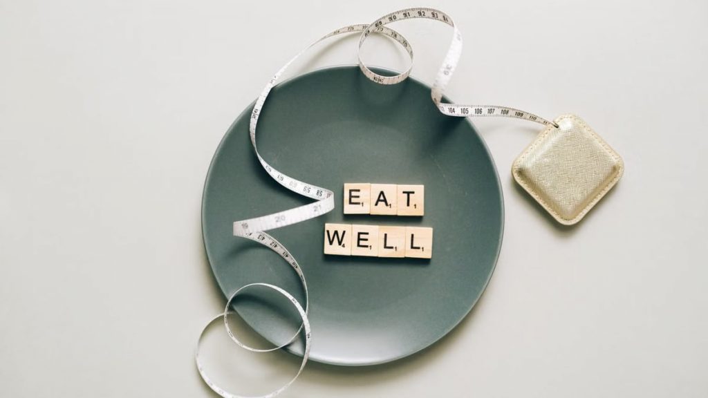 Flat lay of scrabble tiles spelling 'eat well' on a plate with a tape measure, promoting healthy eating.
