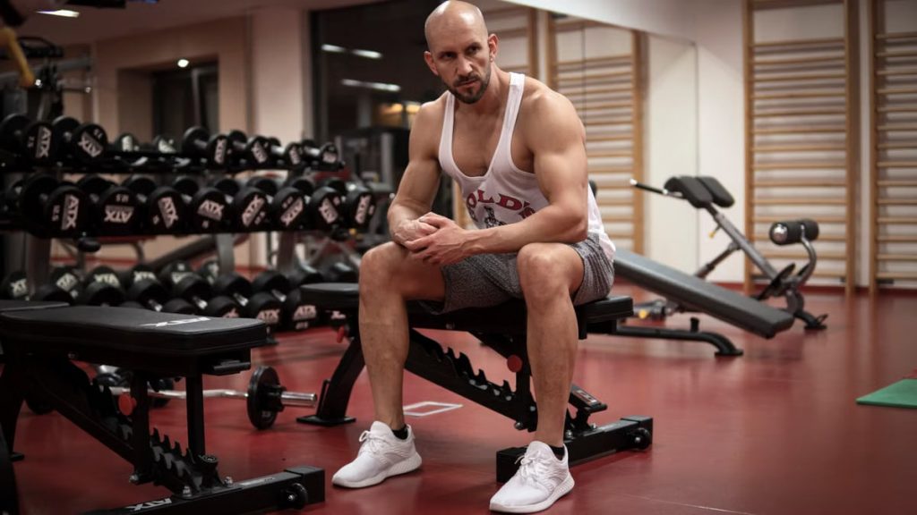 Strong, determined man in white tank top sitting in gym, showcasing fitness and strength.